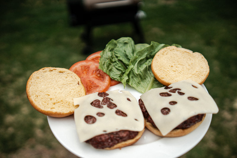 Two wagyu burgers with cheese, tomatoes, and lettuce on a white plate outdoors.